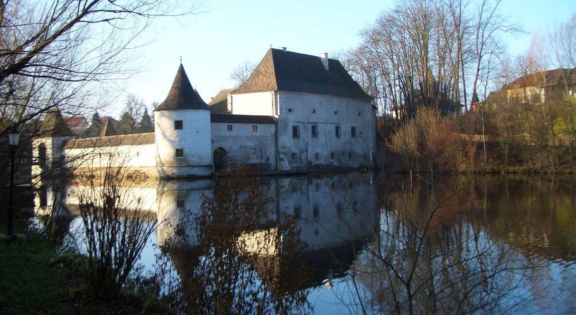 A historic building with towers is reflected in a calm body of water surrounded by bare trees.