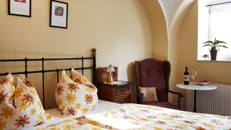 Cozy guest room with floral bedding, armchair, table with wine and fruit, and plants on the windowsill.
