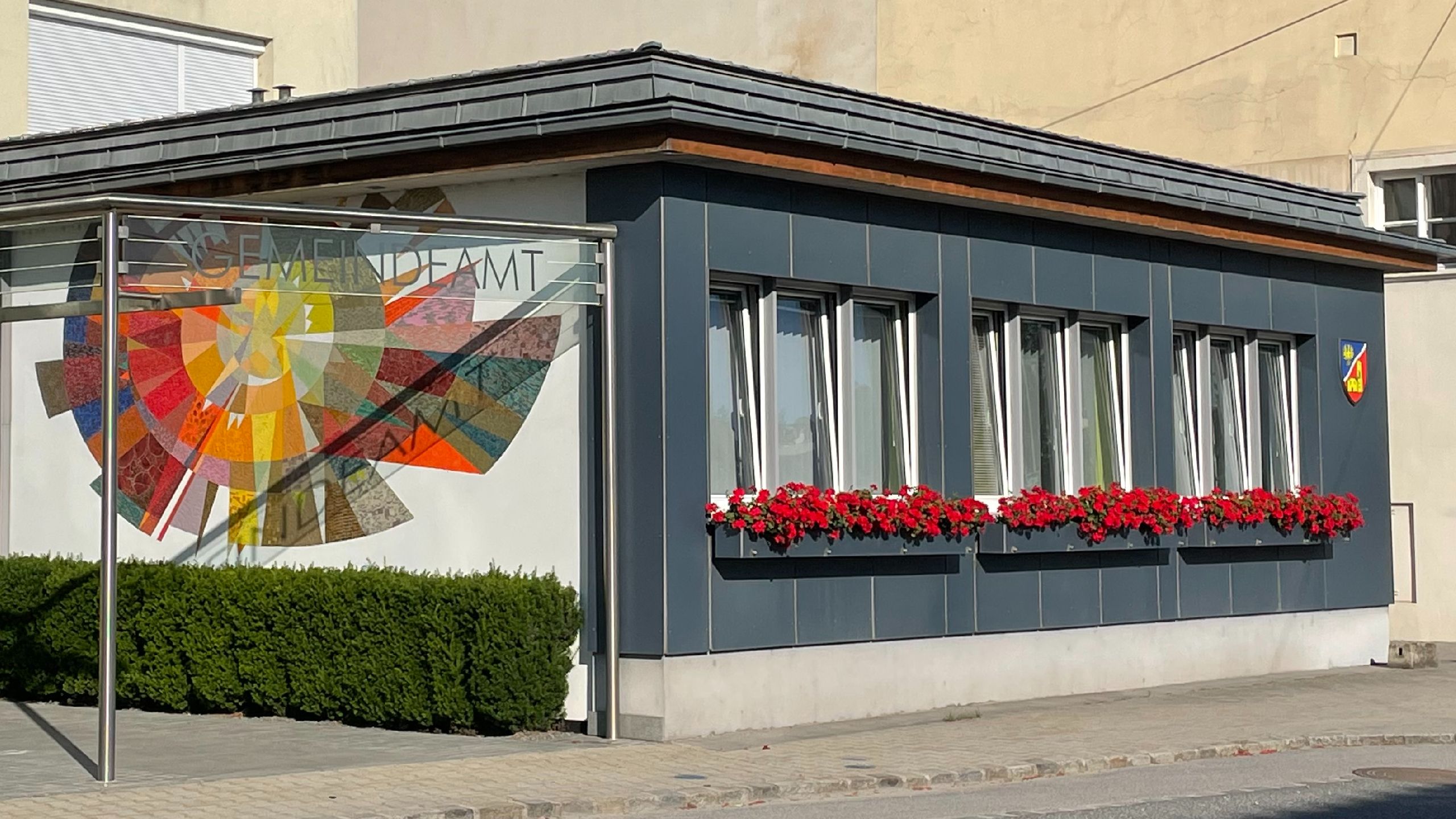 Town hall with a modern facade, a colorful stained-glass window, red flower boxes, and a coat of arms on the wall.