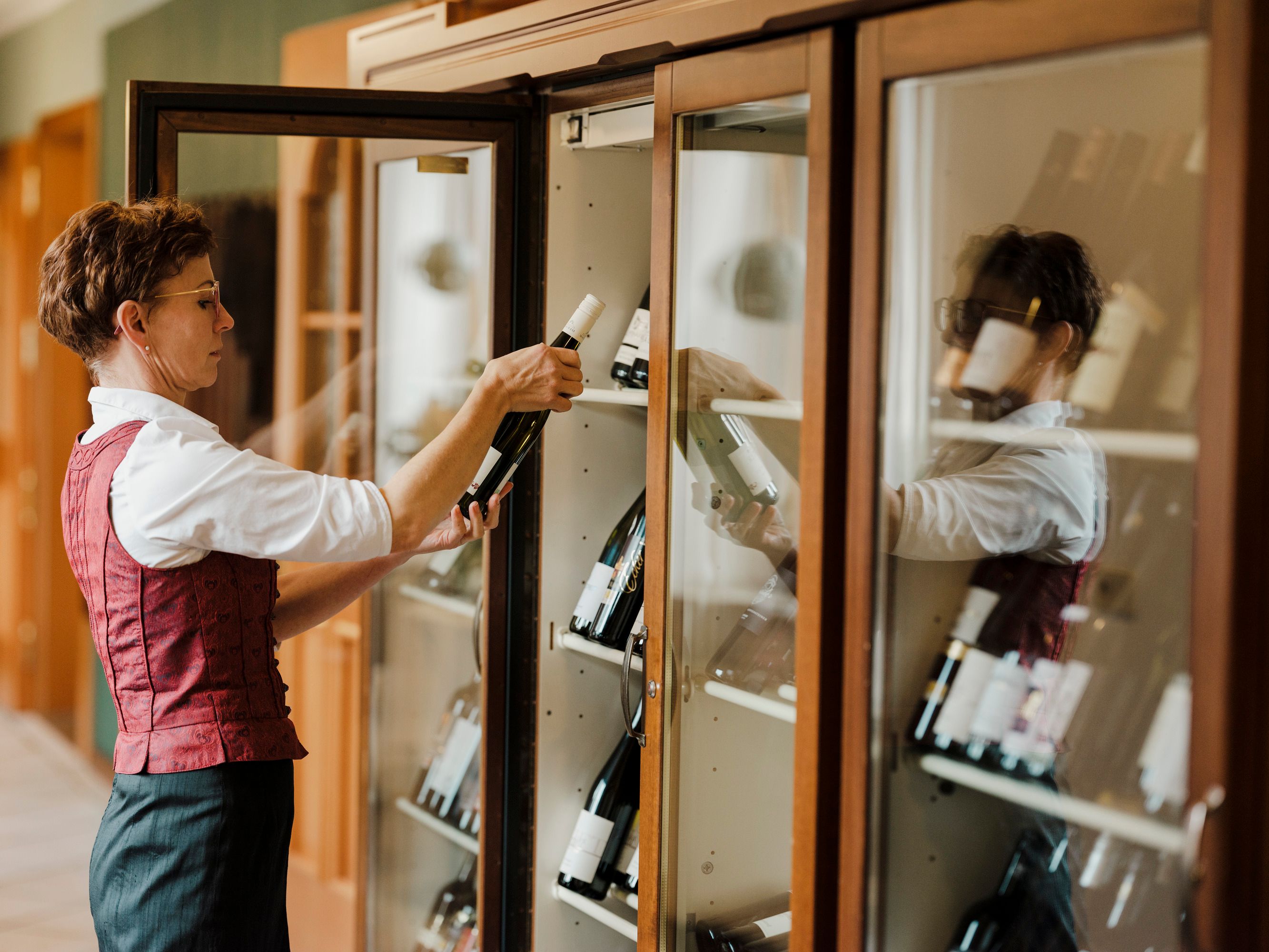 Person in a red vest selects a bottle of wine from a wine cabinet.