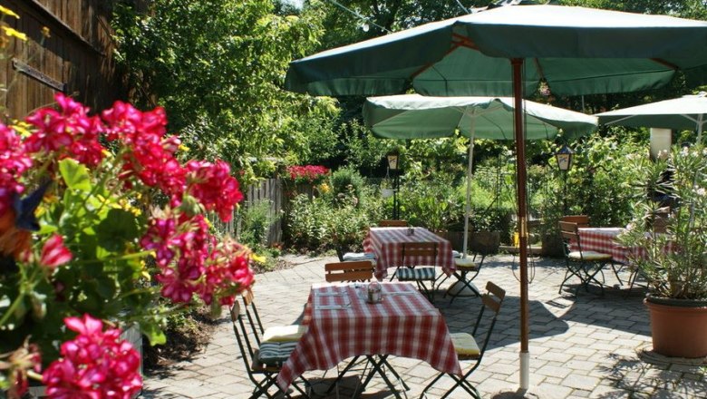 Guest garden, © zVg A cozy guest garden with red and white checkered tablecloths, green parasols and blooming flowers in the foreground.