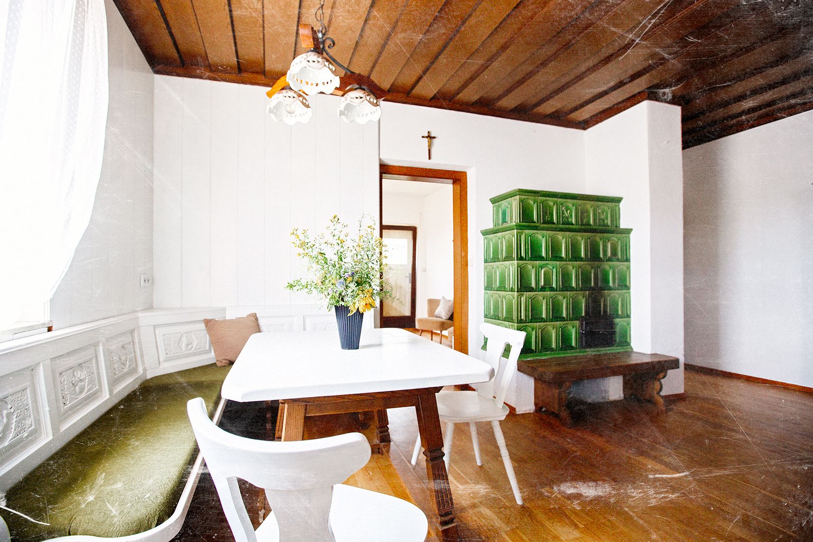 A cozy room with a wooden floor, a white table, white chairs, and a green tiled stove.