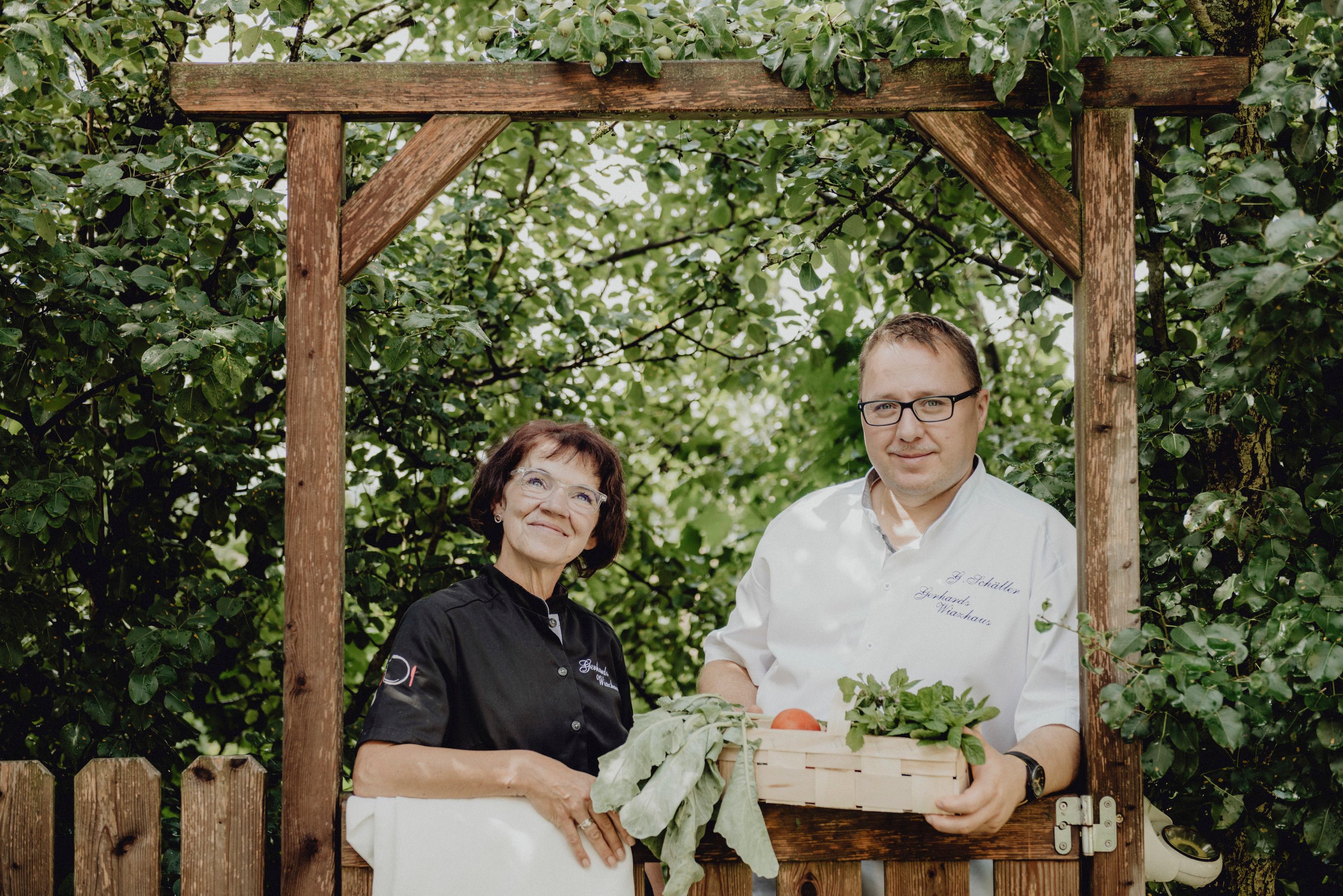 Two people are standing under a wooden arch in the garden, a woman in black clothes and a man in a white shirt with a basket full of vegetables.