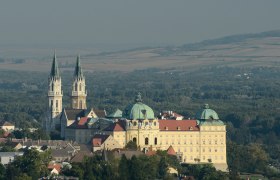 Aerial view of Stift Klosterneuburg Abbey with two towers and a dome, surrounded by forest and fields.