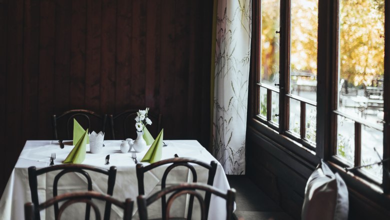 A table in an inn with wooden chairs, white tablecloths and green napkins, next to a window with curtains.