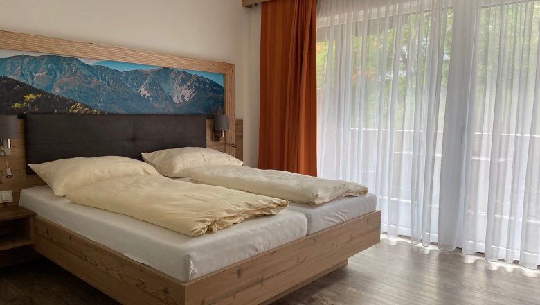 Modern double room with large bed, mountain picture on the wall and large windows with curtains.