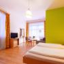 Bright hotel room with double bed, green accent on the wall, TV, seating area and large windows with curtains.