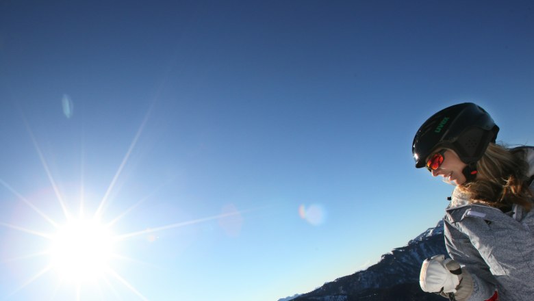 A person wearing a helmet and ski goggles stands in front of a sunny, clear sky in a mountain landscape.