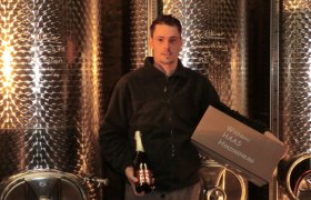 A man stands in front of large stainless steel tanks holding a bottle of sparkling wine and a crate labeled 'Weinbau Haas Herzogenburg'.