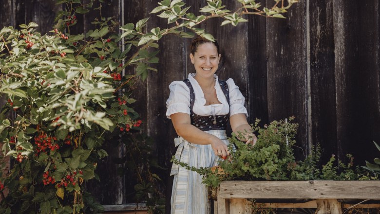 Woman in traditional dress stands smiling next to a bed of herbs in front of a wooden wall.