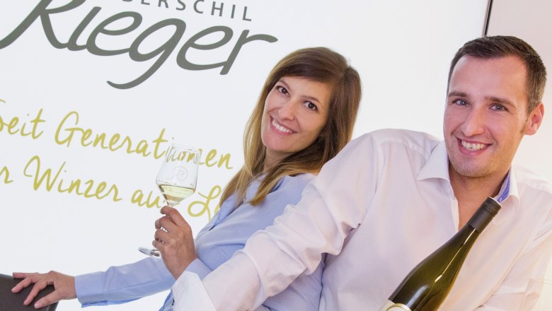 Two people are smiling and holding bottles of wine in front of a sign that reads 'Oberschil Rieger'.