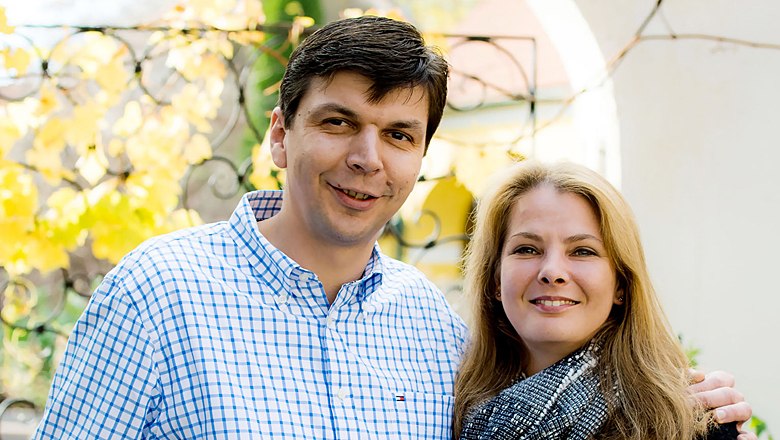 A man and a woman smile at the camera, autumn leaves in the background.