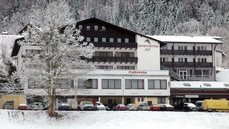 Sporthotel am Ötscher, © Iby & Bogi Matayas Touristik GmbH A snow-covered hotel building with the inscription 'Sporthotel' and 'Cafeteria', surrounded by snow-covered trees and parked cars.
