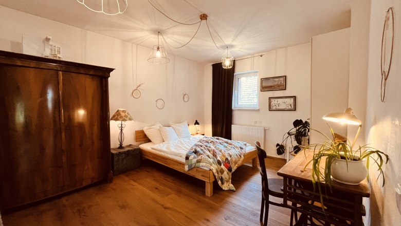 Bedroom, © Elisabeth Fürlinger Cozy bedroom with wooden floor, double bed, antique wardrobe, desk with lamp and plant, as well as decorative elements on the walls.