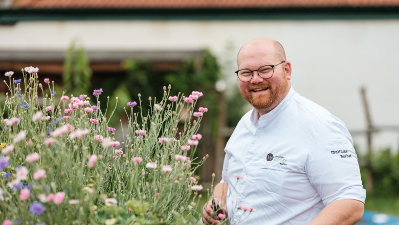 A smiling man in chef's clothes stands next to blooming flowers outside.