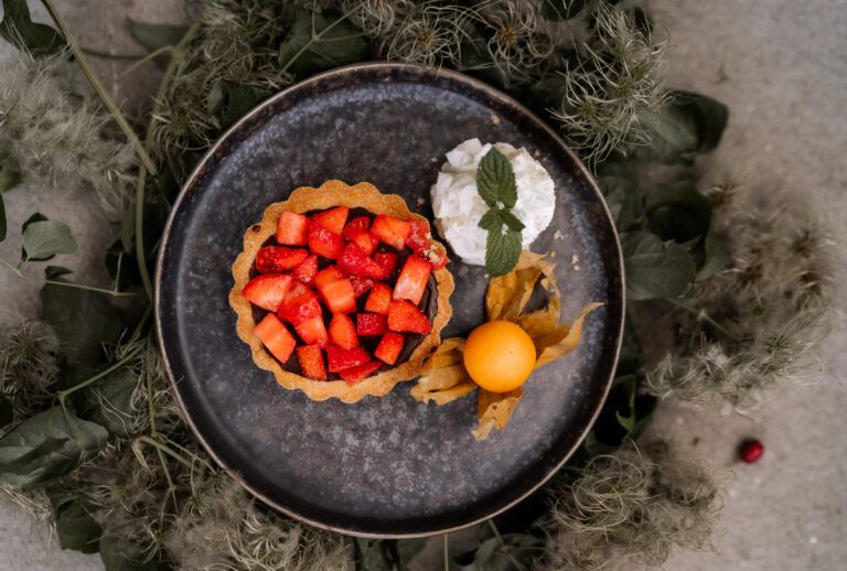A plate of strawberry tarts, cream and physalis, surrounded by green leaves.