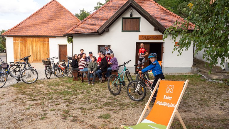 Group of people sitting in front of a building with bicycles, a child stands next to them with a bicycle. A deck chair with an inscription is in the foreground.