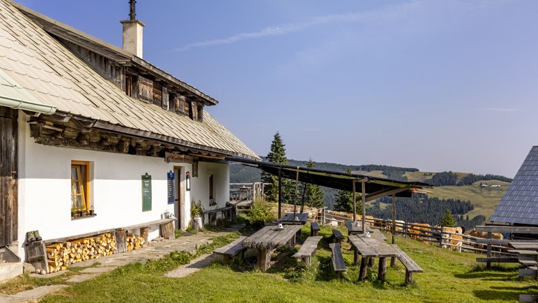 Traditional mountain hut with wooden tables and benches outside, surrounded by green meadows and wooded hills under a blue sky.