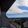View from a balcony of a landscape with houses, trees and hills under a blue sky with clouds.