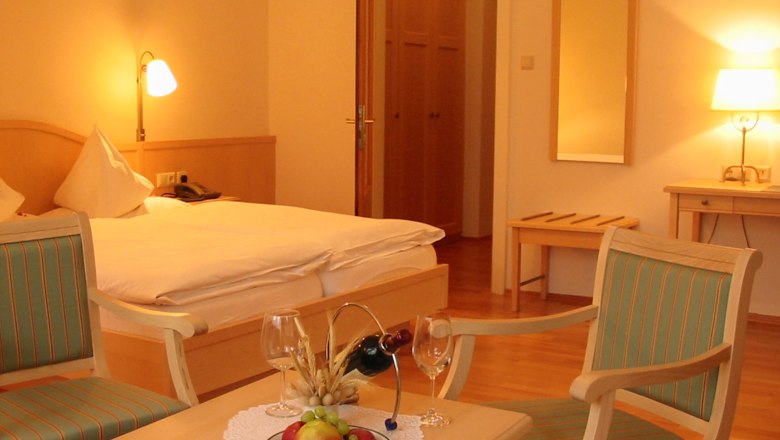 A cozy hotel room with a double bed, two chairs, a table with fruit and wine, and warm light.