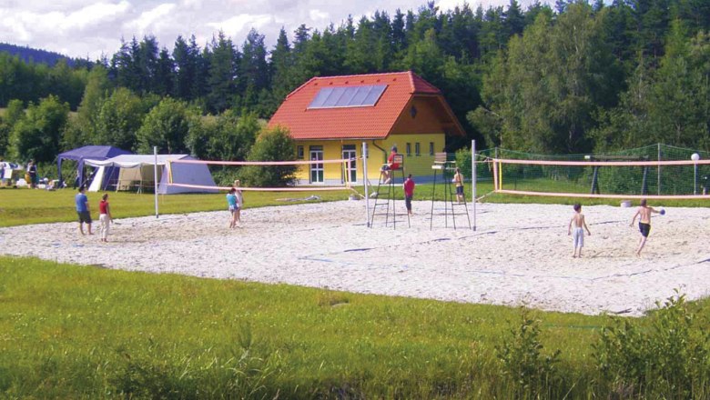 People play volleyball on a sand court in front of a yellow building with a red roof, surrounded by trees.