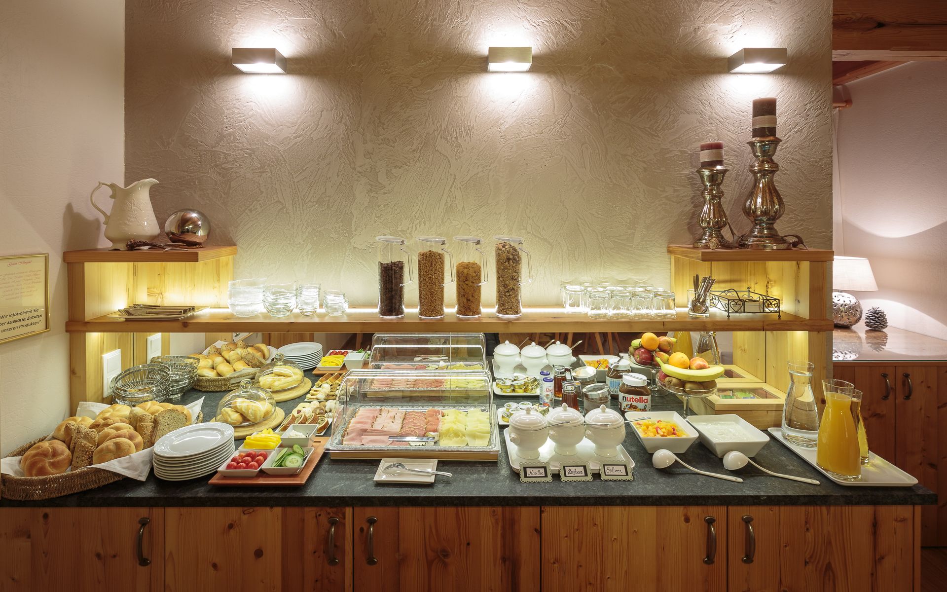 A breakfast buffet with bread rolls, cold cuts, muesli, fruit and drinks on a wooden counter.