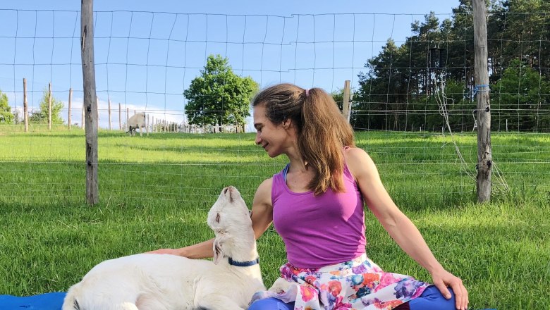 Woman in colorful clothes sits in a meadow next to a goat.