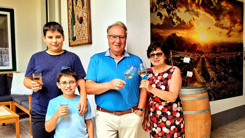 A family stands in front of a wine barrel and a picture of vineyards, holding glasses and smiling at the camera.