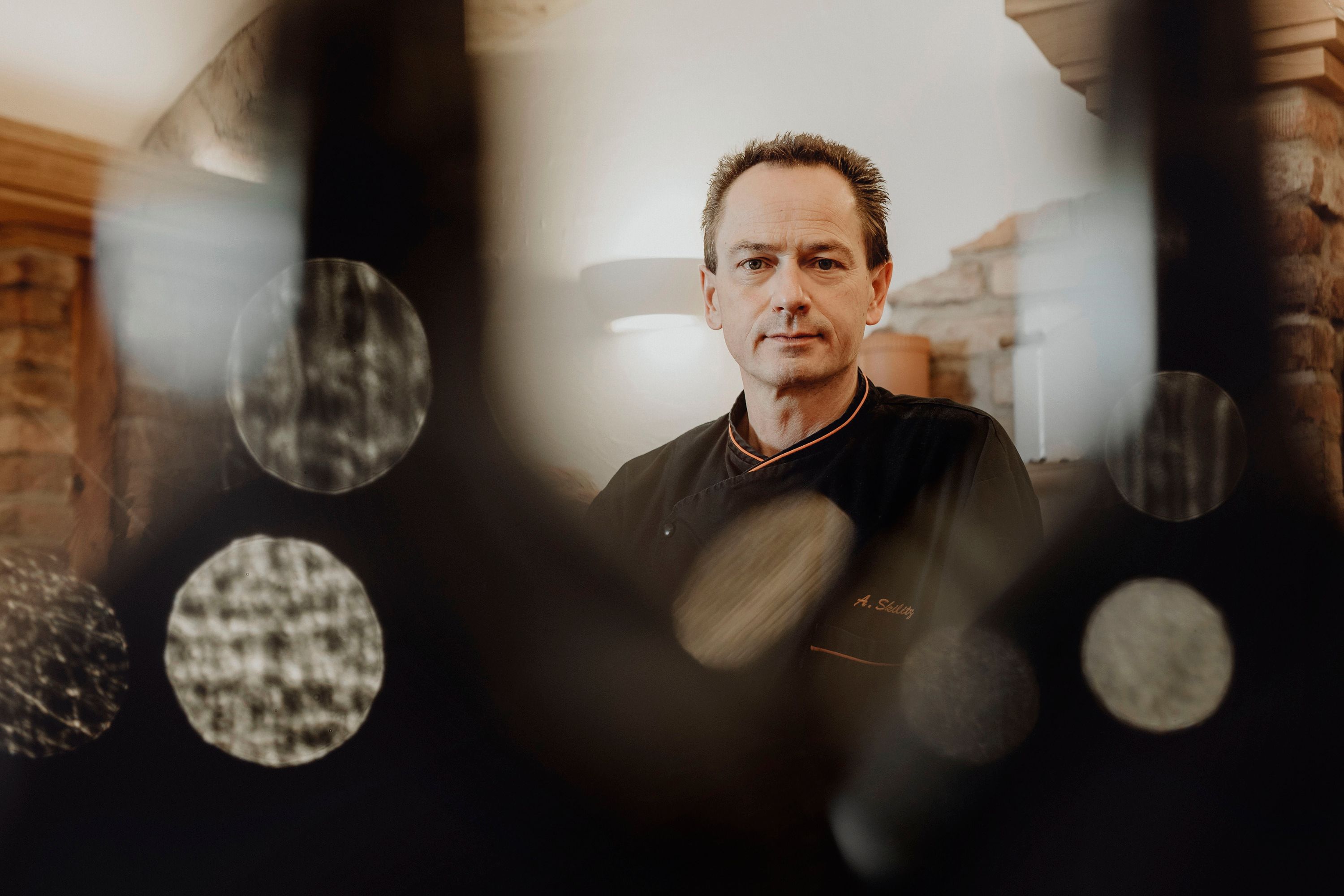 A man in black chef's clothes stands in a rustic room, surrounded by blurred lighting effects.