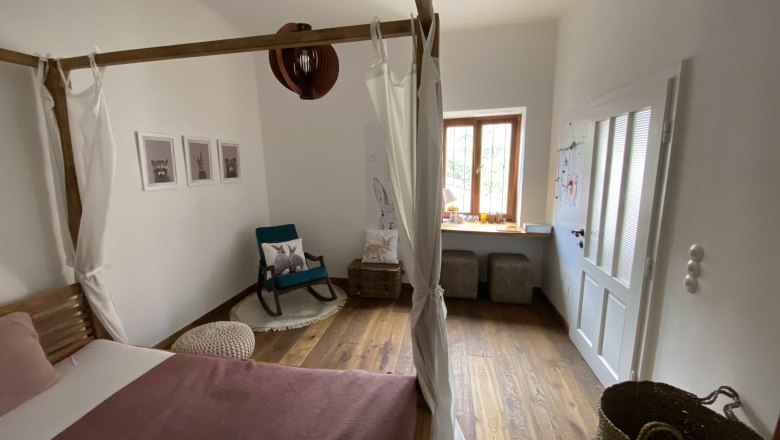 Cozy bedroom with four-poster bed, rocking chair and window seat. Wooden floor and light-colored walls.