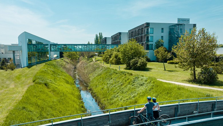 Two cyclists on a bridge in front of the Therme Laa & Hotel.