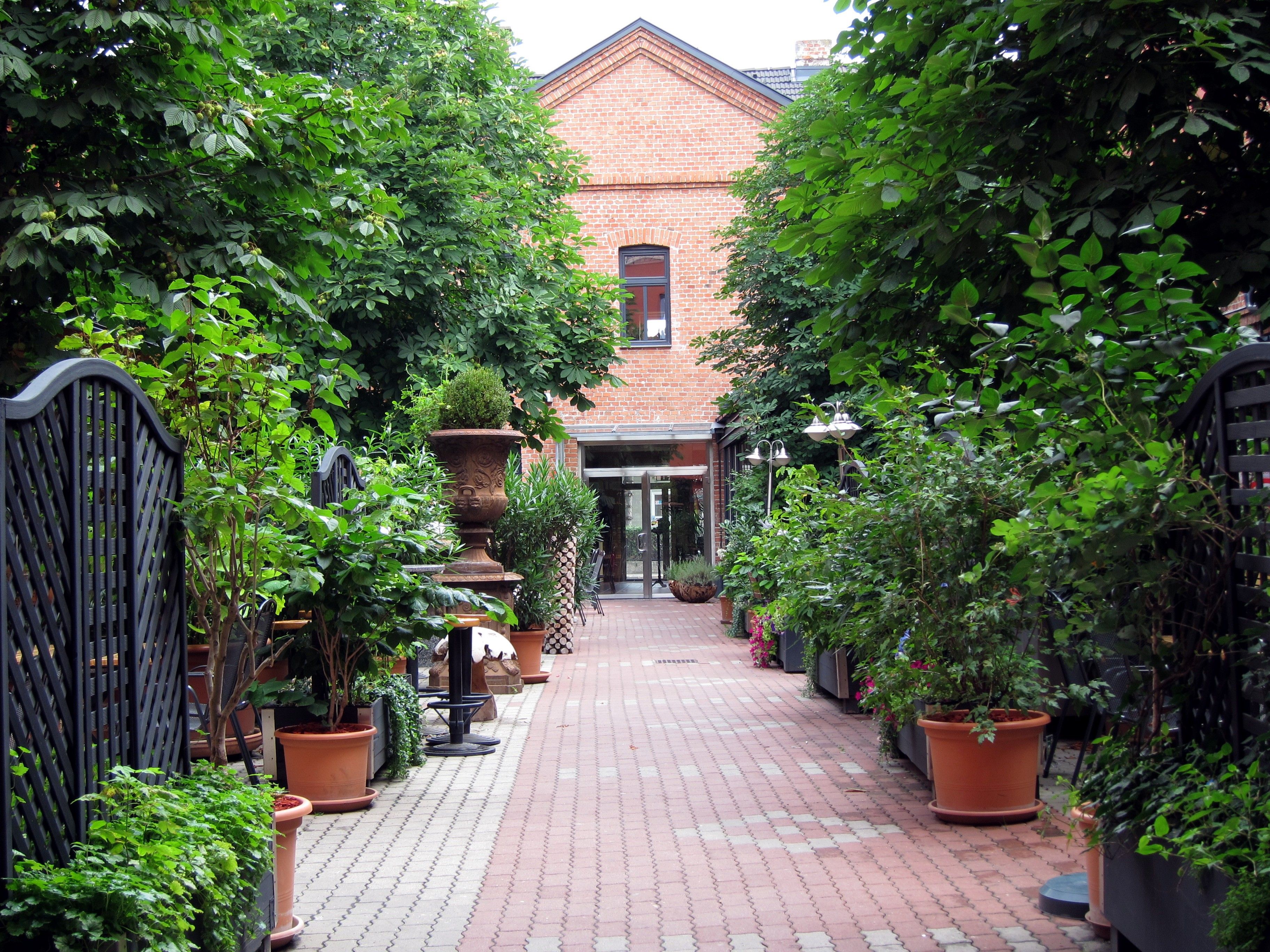 A paved courtyard with plants in pots and a brick building in the background.