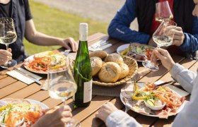 People enjoy wine and cold food outdoors at a wooden table.