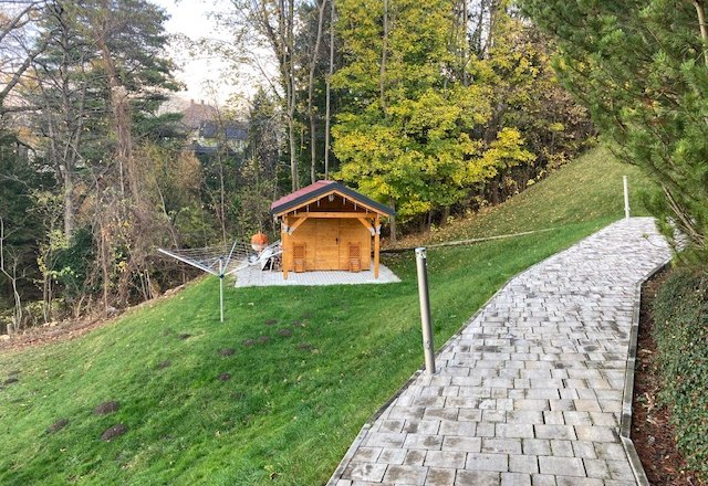 A garden with a small wooden house, surrounded by trees and a paved path.