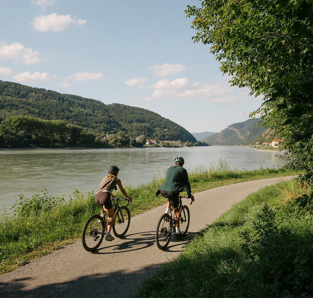 Zwei Radfahrende auf einem asphaltierten Uferweg entlang der Donau; gegenüber liegen bewaldete Hügel, rechts spenden Bäume Schatten.