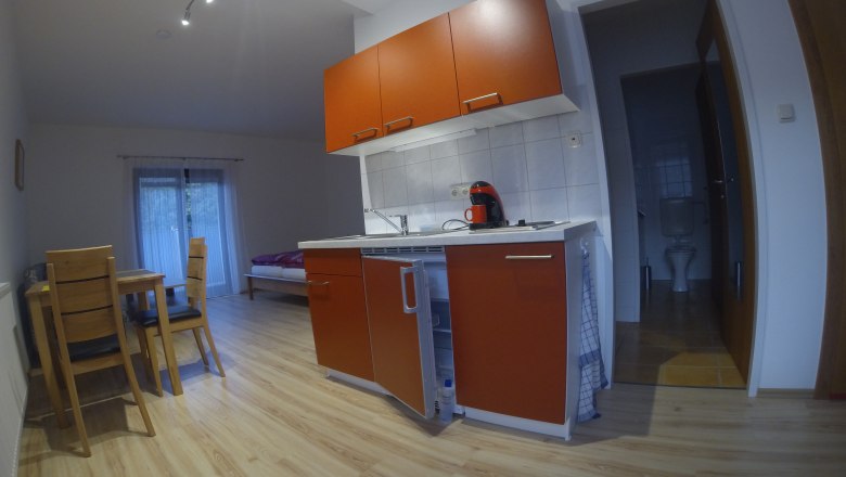 Small kitchen in an apartment with red cupboards, dining table and chairs, bed in the background and bathroom on the right.