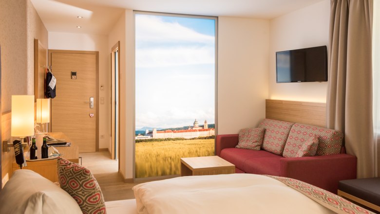 Modern hotel room with double bed, red sofa, flat screen TV and large window with landscape view.