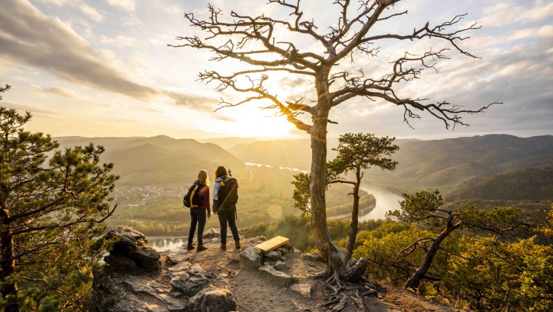 Two people stand on a rock with a view of a river landscape at sunset.