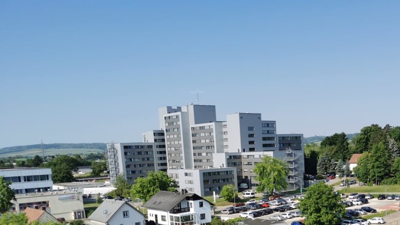 Multi-storey gray building with surrounding trees and cars in a parking lot, blue sky in the background.