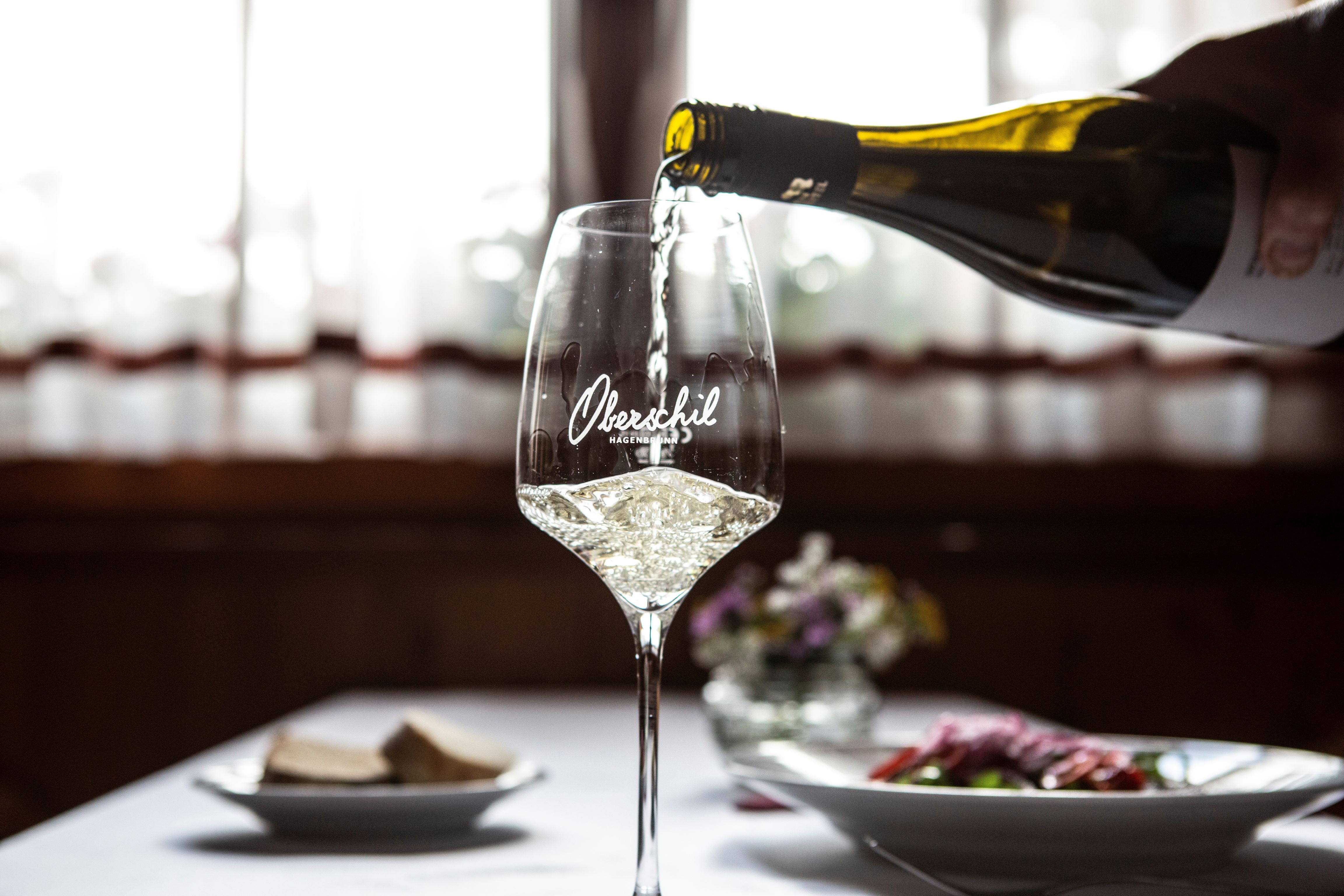 A glass of white wine is poured, a table laid with flowers in the background.