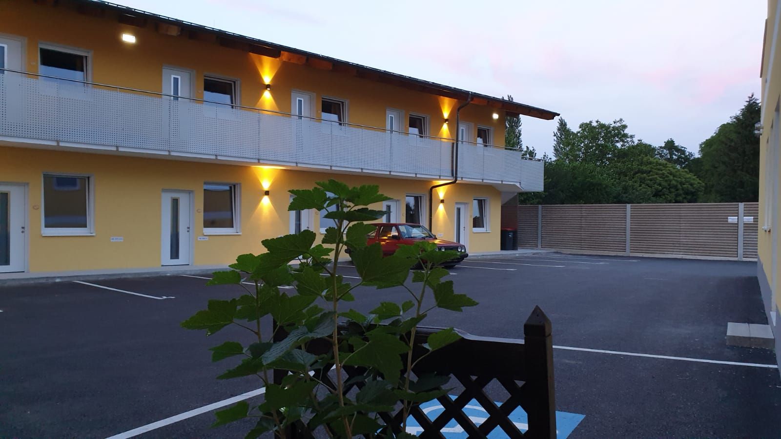 Exterior view of a two-story building with yellow facade and illuminated balcony, in front of it a parked red car and plants in the foreground.