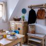 Cozy breakfast corner with wooden table, dishes, yellow cups, chest of drawers, jacket, rucksack and flowers on a cupboard.
