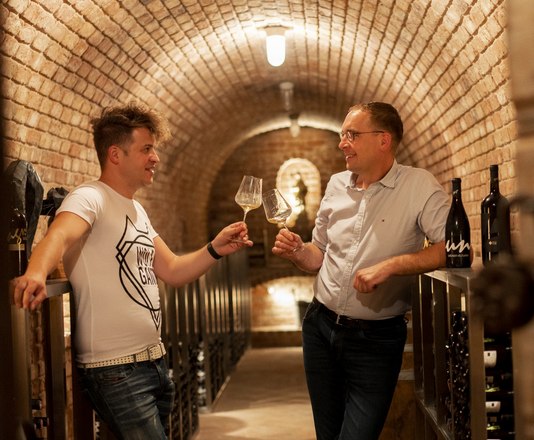 Two men are standing in a wine cellar, clinking glasses of wine.