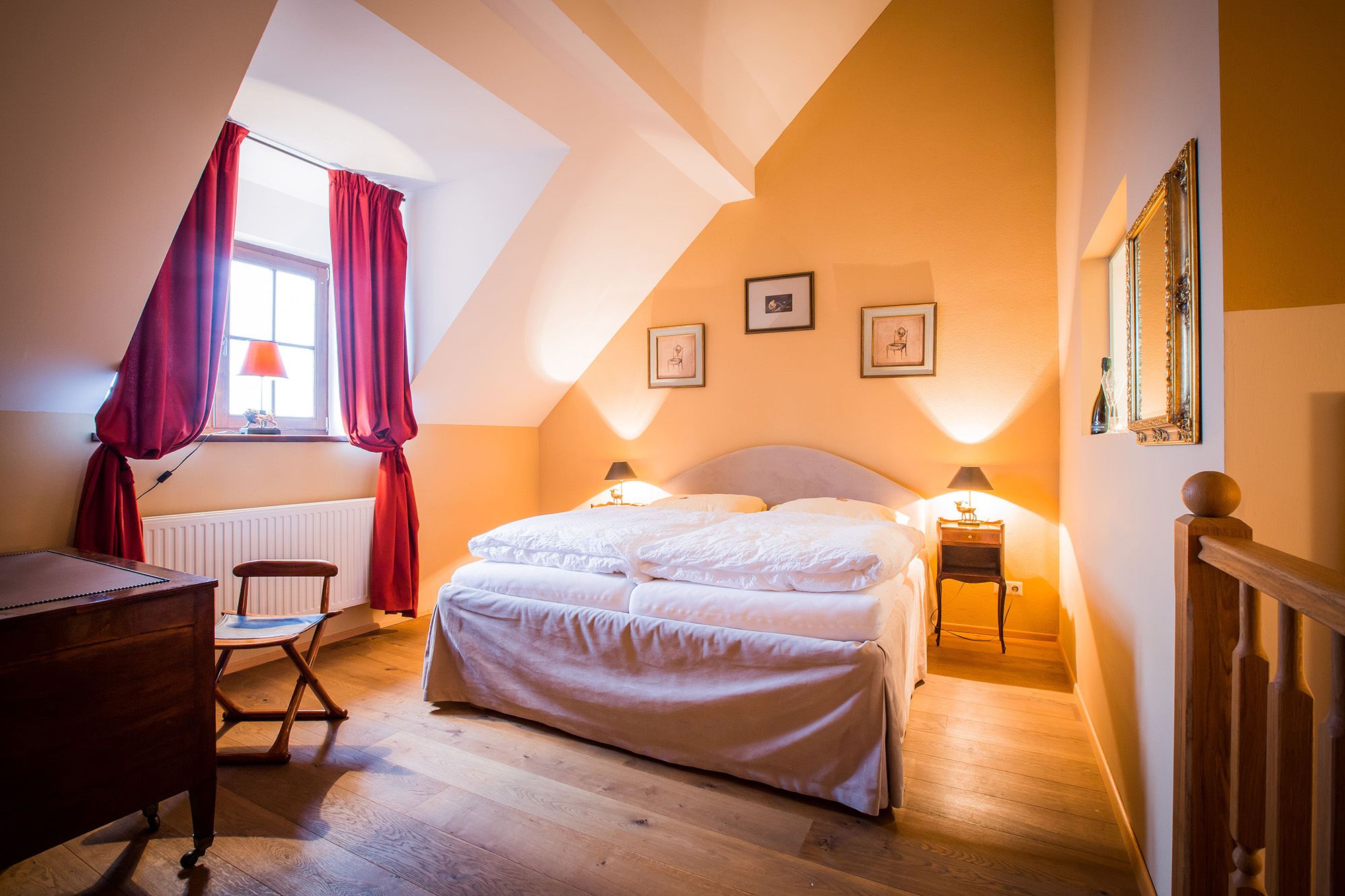 A cosy double room with a large bed, red curtains and warm colors. Pictures hang on the wall and a window lets in natural light.