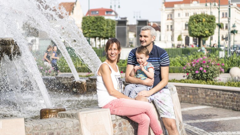 A family sits by a fountain in a town with historic buildings in the background.