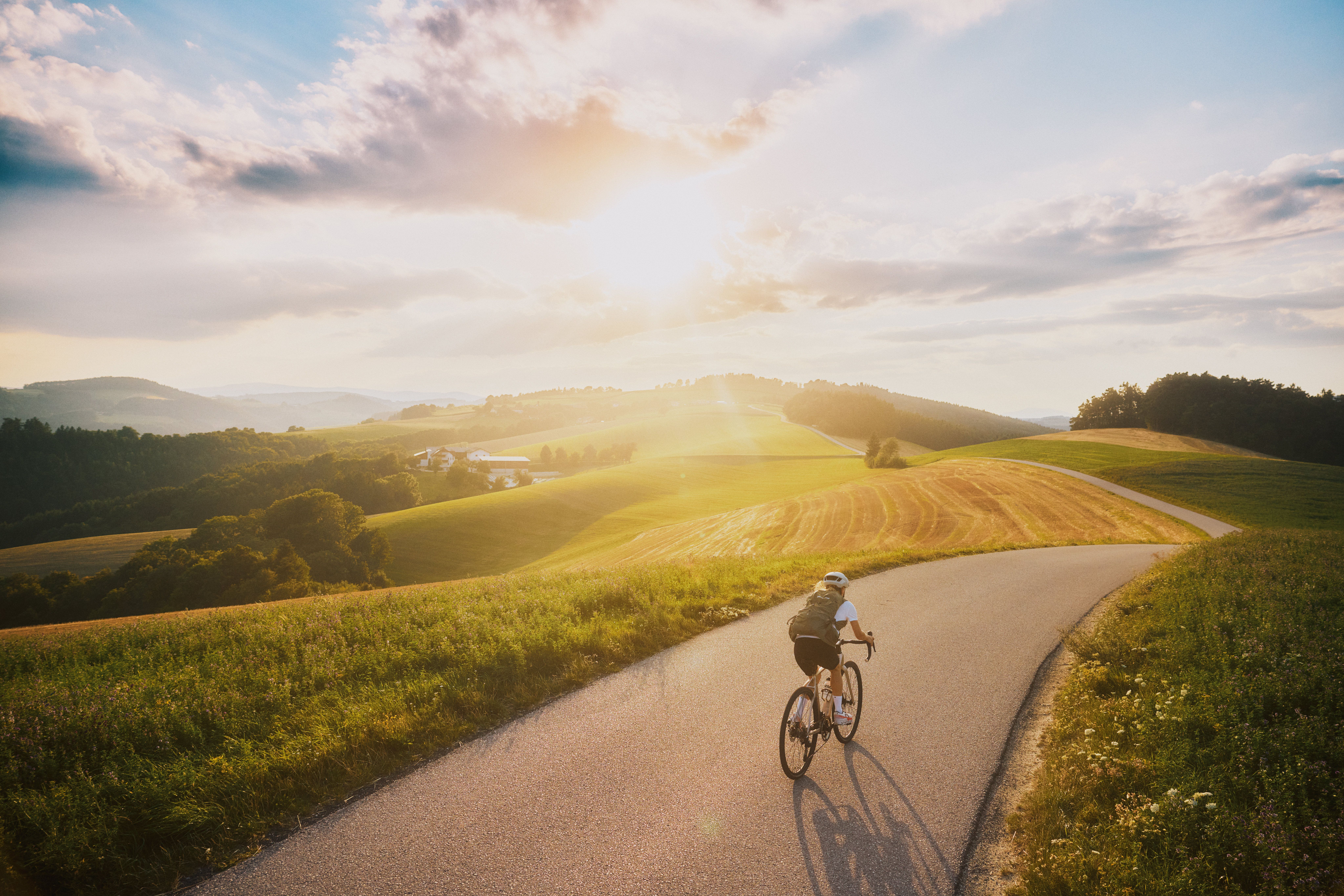 A cyclist rides along a winding country road through a hilly landscape at sunset.