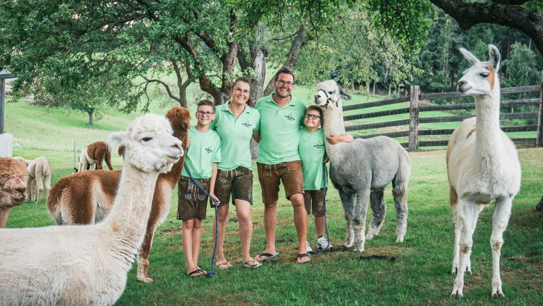 A family in green T-shirts stands in a meadow with several alpacas.