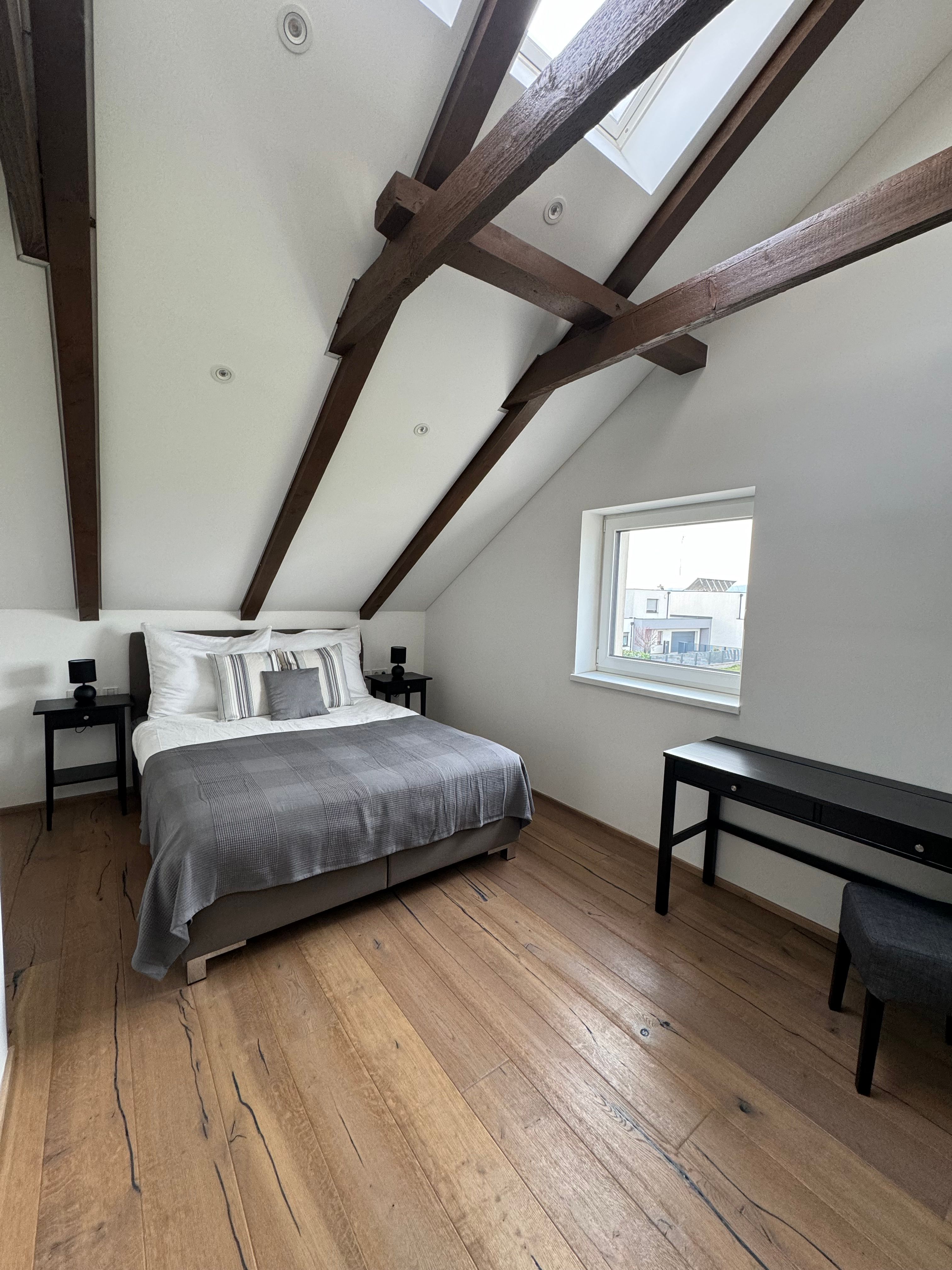 Modern bedroom with wooden beamed ceiling, double bed, wooden floor and window.