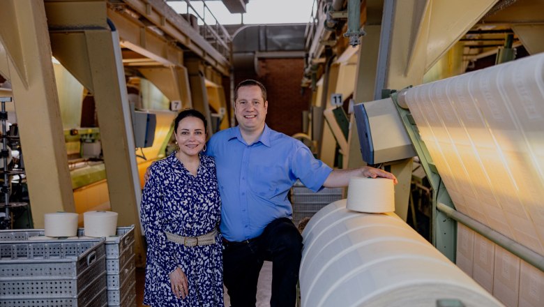 Two people stand next to machines and rolls of fabric in a textile factory.