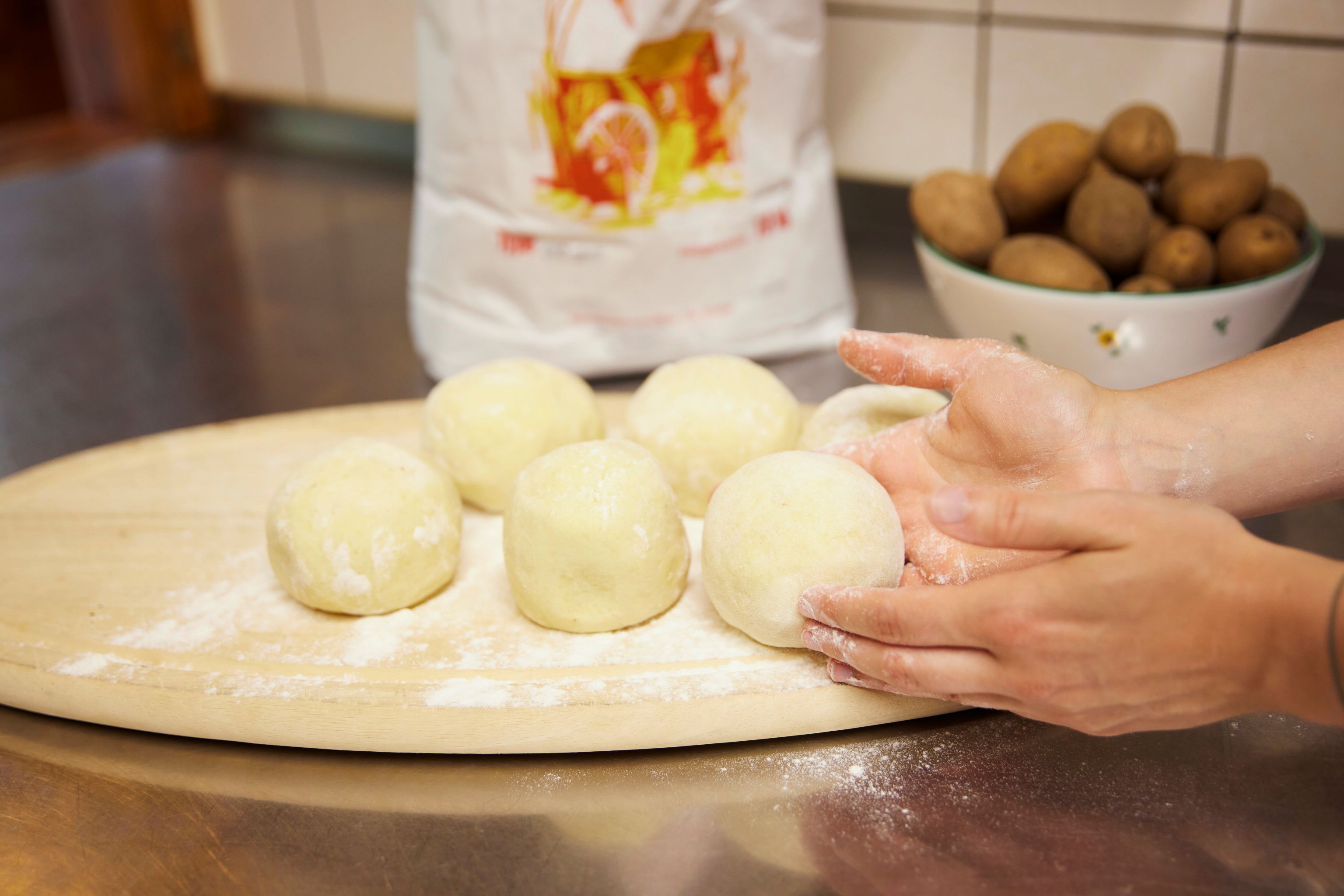 Hands form dumpling dough on a floured wooden board, with a bowl of potatoes in the background.